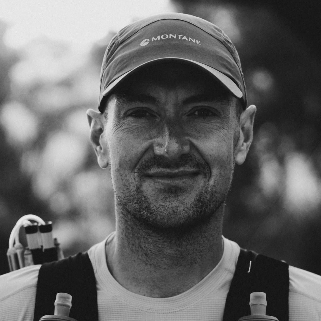 A black and white close-up portrait of a man wearing a Montane baseball cap, a white athletic shirt, and hydration straps over his shoulders, with a blurred outdoor background.