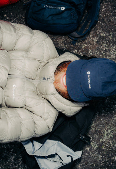 A person lying on a rocky ground wearing a light beige puffer jacket, a navy blue baseball cap with Montane logo, and a dark blue backpack is visible in the background.
