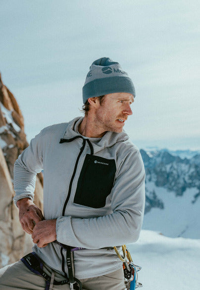 A male climber wearing a light gray hooded fleece jacket with a black chest pocket, a blue beanie, and climbing harness gear, posed outdoors against a snowy mountain background.