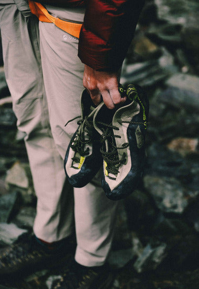 A person outdoors on a rocky surface holds a pair of climbing shoes in one hand. The shoes are dark gray and off-white with black laces and small neon yellow-green accents.