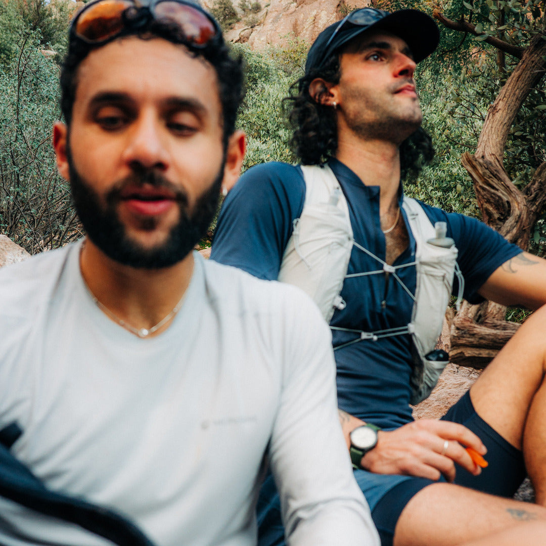 Two men outdoors in a rocky, wooded setting. The man in the foreground wears a white long-sleeve shirt and has sunglasses on his head.