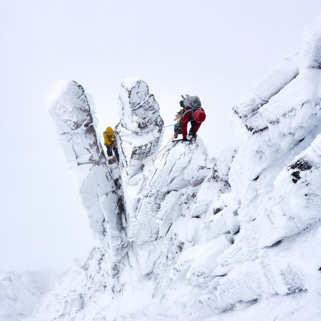 Two mountaineers climb a steep, snow-covered rock formation in a snowy outdoor environment. One climber wears a bright red jacket and dark pants, while the other is further up the slope in a yellow jacket.
